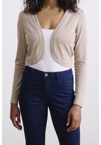 Beige, kurz geschnittene Strickjacke mit langen Ärmeln, weichem Stricktextur und abgerundetem, offenem Vorderteil. Kombiniert mit dunkelblauen, figurbetonten Jeans.