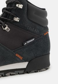 Bota de senderismo de ante negro con una superficie texturizada, con herrajes plateados, un detalle de costura en naranja y una suela resistente con un patrón de camuflaje.