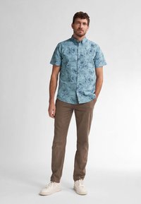 Camisa de botones de manga corta en azul claro con patrón floral, combinada con pantalones beige y zapatillas blancas. La camisa tiene un cuello clásico y un corte regular.