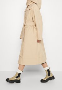 Personne marchant portant un long manteau beige avec ceinture, des chaussettes blanches et des bottines beiges et noires à semelle épaisse et brillante.