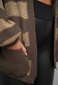 Brun og kremfarget strikket cardigan med løs passform. Den har en lomme og teksturert mønster, kombinert med svarte tettsittende leggings.