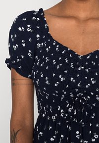 Navy floral dress with short puff sleeves. Features gathered bodice with a tie detail and small white flowers against a dark background.