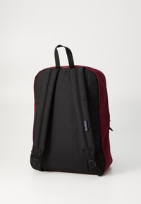 Un sac à dos avec une partie supérieure bordeaux et une section inférieure noire, doté de bretelles rembourrées, d'une poignée sur le dessus et d'un petit patch de logo sur le côté.