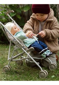 Kind gekleed in een beige fuzzy jack en een donkere beanie, die een pop in een grijze kinderwagen vastmaakt. De pop heeft lang blond haar en draagt een groene outfit.