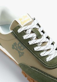 Basket verte olive avec un mélange de textures lisses et en daim, lacets blancs et un logo de tête de mort avec des os croisés sur le côté.