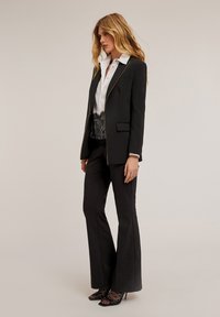 Blazer negro ajustado con detalles de cremallera dorada, combinado con una camisa blanca y pantalones negros. Presenta un borde de encaje a lo largo de la cintura y tacones abiertos.