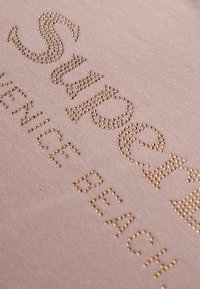 Camisa rosa claro con tachuelas doradas en relieve que dicen "SOPORTE VENICE BEACH". Tejido suave con una textura lisa y un diseño de letras claro.