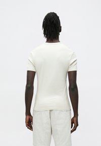 Homem de pele escura e dreadlocks, vestido com uma camisa bege de mangas curtas e calções de cor clara, de costas para um fundo simples.