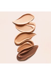 Cinq échantillons de fond de teint liquide dans différentes nuances de beige et de marron, superposés verticalement sur un fond clair.