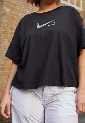 Personne portant un t-shirt noir ample avec un swoosh Nike argenté et un slogan, associé à un pantalon blanc, debout devant un mur en briques.