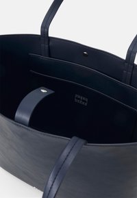 Marinblå läder toteväska med två långa handtag, en strukturerad utsida och en präglad logotyp på en ficka inuti. Slät inredning.