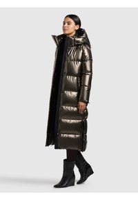 Duga jakna na preklop u metalik tamno smeđoj boji, s kapuljačom, patentnim zatvaračem i kopčama te prošivenim dijelovima; nošena preko crne majice i hlača.