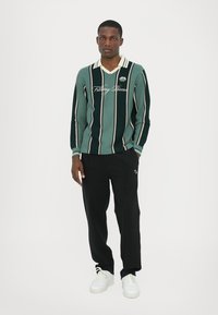 Grüner und marineblauer gestreifter Strickpullover mit V-Ausschnitt, versehen mit weißen Akzenten und Logo. Kombiniert mit schwarzen Hosen und weißen Sneakers.