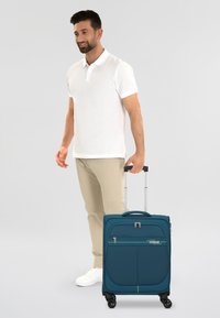 Teal rollenkoffer met een harde schaal, dubbele rits voorvak, uitschuifbare handgreep en vier wielen voor mobiliteit.