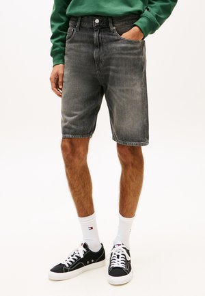 Homme portant un short en jean noir délavé, des chaussettes blanches montantes, des baskets noires et une chemise verte à manches longues, debout avec une main dans la poche.