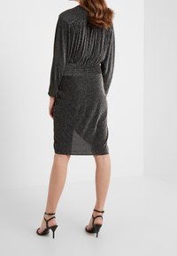 Czarna plisowana sukienka z metalicznymi nitkami, dopasowana sylwetka, długie rękawy i długość do kolan; zestawiona z czarnymi sandałami na cienkim obcasie.