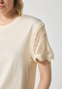 Cream cotton t-shirt featuring a delicate, crocheted lace sleeve. The lace is light beige with scalloped edges, adding a textured detail.