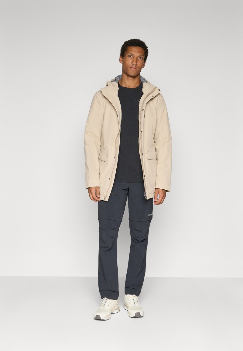 Beige parka met een capuchon, ritssluiting aan de voorkant en twee zijzakken, gecombineerd met donkergrijze cargobroek en lichte schoenen. Glad textuur.
