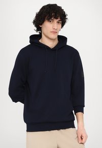 Felpa blu navy con tasca frontale, coulisse regolabile e polsini a coste. Realizzata in tessuto morbido, presenta una toppa testurizzata sulla manica.