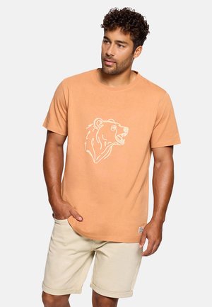Hombre con cabello rizado que lleva una camiseta naranja con un gráfico de cabeza de oso blanco y pantalones cortos beige, de pie con una mano en el bolsillo.