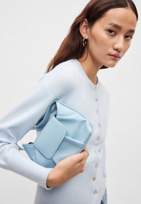 Τσάντα χειρός από ανοιχτό γαλάζιο δέρμα με δομημένο σχεδιασμό, που διαθέτει εμπρόσθιο καπάκι, απαλής υφής και ρυθμιζόμενο λουράκι, κρατημένη πάνω σε χρώμα απαλού γαλάζιου πουλόβερ.