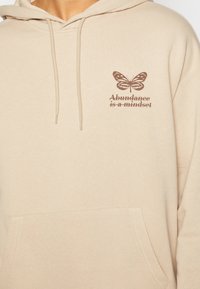 Sudadera con capucha beige con un bolsillo frontal tipo canguro, que presenta un gráfico de una mariposa marrón y el texto "La abundancia es una mentalidad" en una fuente moderna.