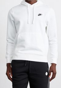 Bílá mikina s kapucí vyrobená z bavlněné směsi, s přední kapsou, šňůrkou na kapuci a černým logem Nike na levé hrudi. Nosí se s černými kalhotami.