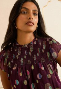 Blusa bordeaux con motivi a pois multicolori scintillanti, dotata di un corpino arricciato e maniche corte con volant. Tessuto morbido e leggero.