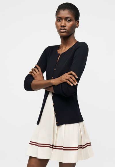 Γυναίκα με σταυρωμένα χέρια που φοράει μαύρο ζακέτα με κουμπιά και λευκή πλισέ φούστα με δύο οριζόντιες καφέ ρίγες κοντά στο τελείωμα.