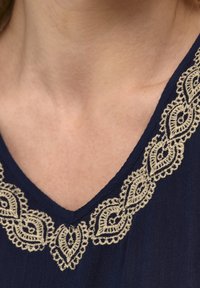 Dunkelblaues Oberteil mit V-Ausschnitt und kunstvoller cremefarbener Stickerei in einem Blättermuster um den Halsauschnitt. Weicher Stoffurtaltet sichtbar.