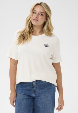 Szőke nő fehér, rövid ujjú ingben, amelyen apró fekete tengeri kagyló minta található, és kék farmernadrágot visel, egy egyszínű háttér előtt áll.