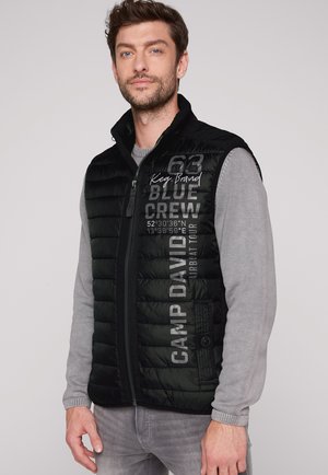 Hombre con barba que lleva un chaleco negro acolchado sobre un suéter gris, el chaleco muestra texto que incluye "Camp David" y "Blue Crew" en el frente.