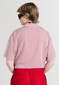 Persona con capelli corti castano chiaro che indossa occhiali da sole neri, una camicia a maniche corte a strisce verticali rosse e bianche e pantaloni rossi a vita alta.