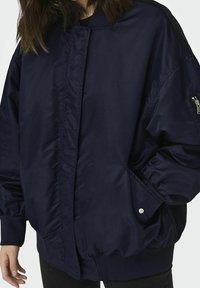 Navy modrá vycpaná bomber bunda s elastickými manžetami a spodním lemem. Má boční kapsy a zipovou kapsu na levém rukávu. Hladký, lesklý materiál.