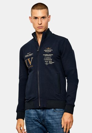 Giacca navy con zip e colletto alto, caratterizzata da dettagli testurizzati, loghi stampati in oro e polsini a coste. Inclusi due taschini laterali.