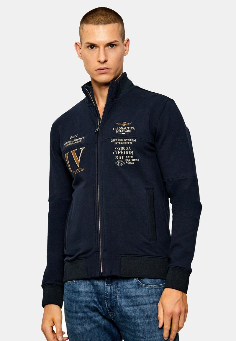 Giacca navy con zip e colletto alto, caratterizzata da dettagli testurizzati, loghi stampati in oro e polsini a coste. Inclusi due taschini laterali.