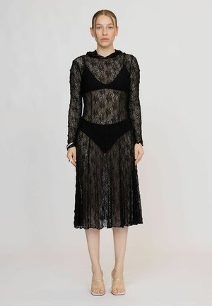Femme portant une robe longue en dentelle noire transparente à manches longues dessus un soutien-gorge noir et des sous-vêtements, debout sur un fond clair avec une expression neutre.