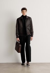 Veste bomber en cuir marron foncé avec deux poches avant, portée sur un col roulé noir. Le mannequin tient un sac en cuir marron et porte des chaussures assorties.