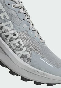 Zapatilla de correr gris con una parte superior de malla, patrones texturizados, acentos reforzados, una suela LIGHTSTRIKE y un logotipo prominente en gris.
