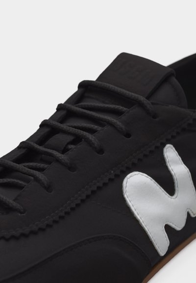 Κοντινό πλάνο ενός μαύρου αθλητικού παπουτσιού με μαύρα κορδόνια και ένα λευκό αφηρημένο λογότυπο στο πλάι, πάνω σε απλό φόντο.