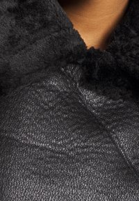 Veste en cuir noir avec un col en shearling doux et texturé. La surface présente un motif de grain naturel, mettant en avant la qualité du matériau.