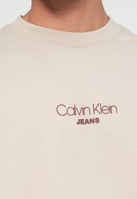 Bežs kokvilnas T-krekls ar apaļu kaklu, ar bordo krāsas "Calvin Klein JEANS" logo uzdrukātu priekšpusē. Gluda tekstūra.