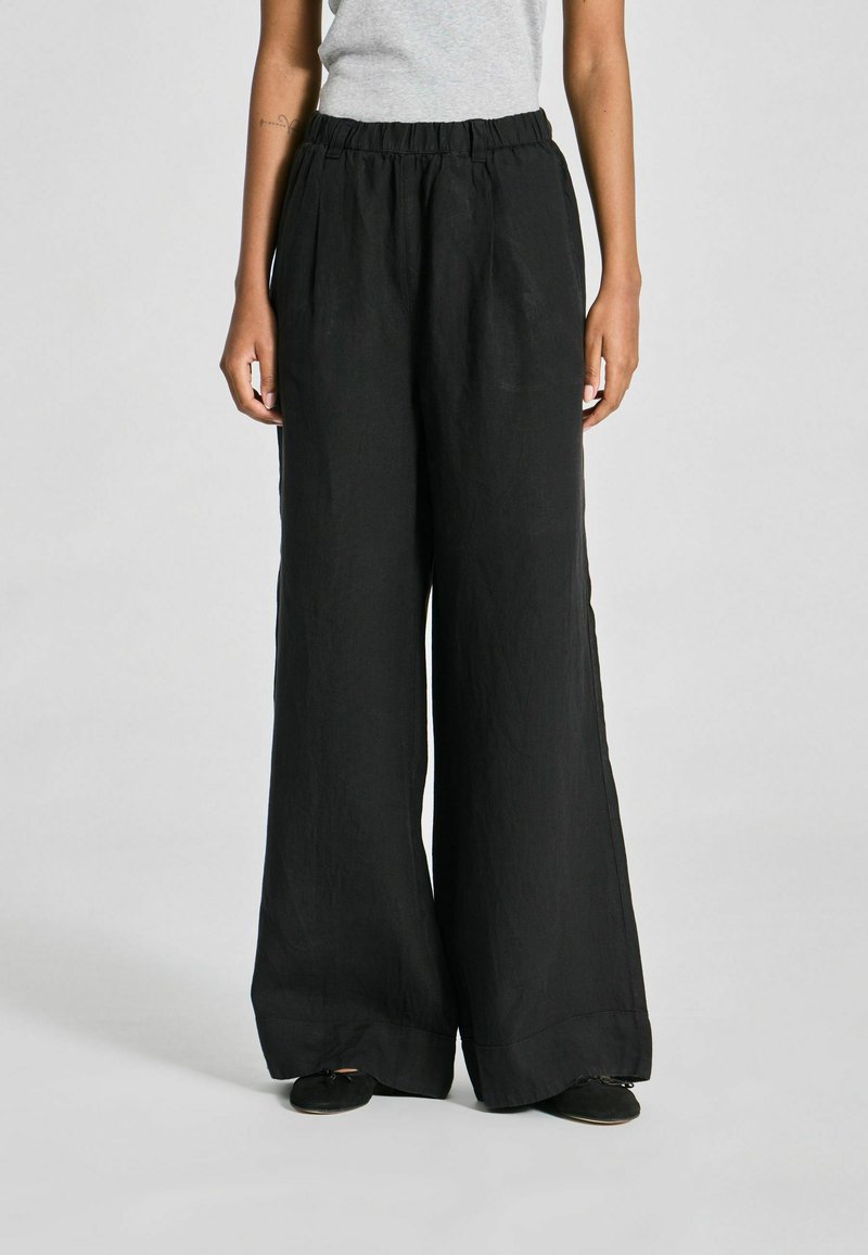 Personne portant un pantalon large noir ample et des chaussures noires, debout devant un fond clair uni.