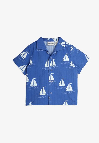Kortermet, blå skjorte med et mønster av hvite seilbåter og bølger. Teksturert stoff med krage og knappelukking.