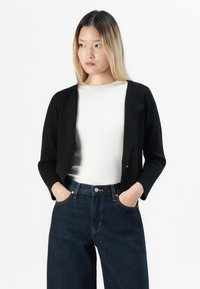Jeune femme aux cheveux blonds raides portant un cardigan noir sur un haut blanc et un jean taille haute bleu foncé, les mains dans les poches, regardant vers la gauche.