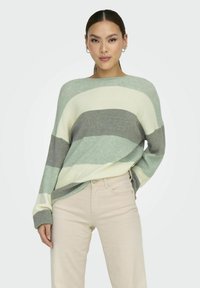 Un pull rayé en vert, gris et crème, à la coupe large et à la texture côtelée. Assorti à un pantalon beige clair pour contraster.