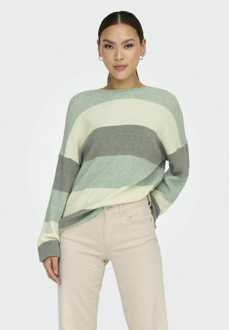 Un pull rayé en vert, gris et crème, à la coupe large et à la texture côtelée. Assorti à un pantalon beige clair pour contraster.