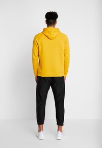 Sudadera con capucha amarilla y un bolsillo tipo canguro, combinada con pantalones negros ajustados y zapatillas blancas, vista desde atrás.