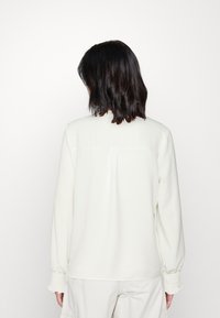 Weiße Bluse mit langen Ärmeln, entspanntem Schnitt, weicher Textur, gerüschten Manschetten und dezenten Rückennähten. Keine sichtbaren Logos oder Muster.