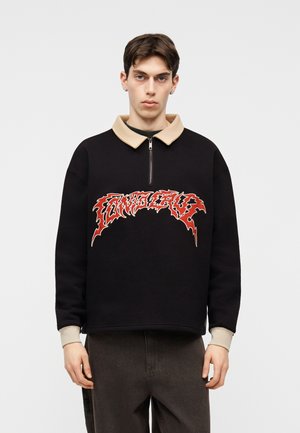 Νεαρός άνδρας με κοντά καστανά μαλλιά, φορώντας μαύρο πουλόβερ με μπεζ γιακά και κόκκινο γραφικό κείμενο στο στήθος, στέκεται μπροστά σε μονόχρωμο φόντο.